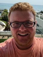 Man in orange shirt wearing glasses smiling in front of a beach front